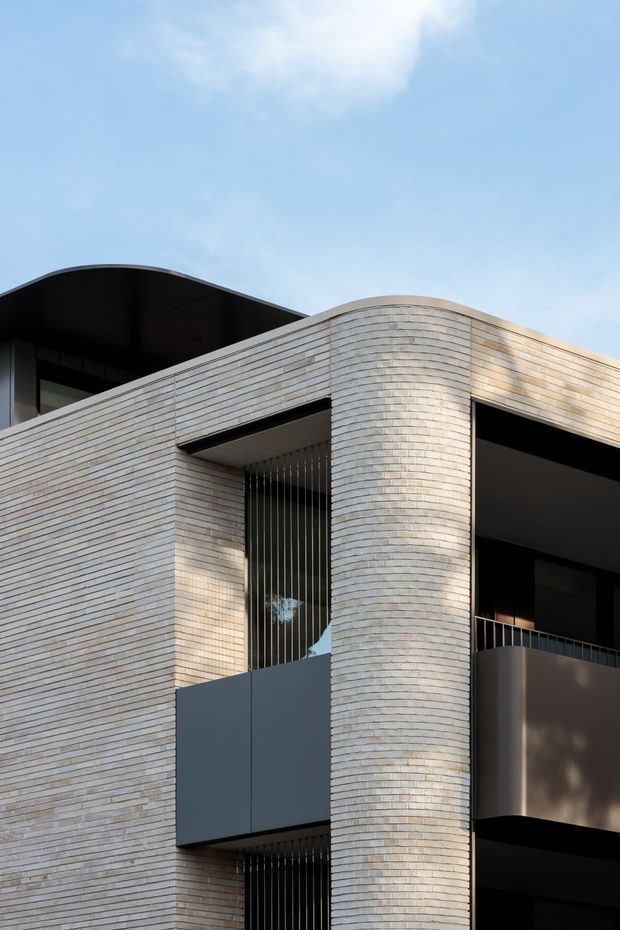 Close-up architectural photograph of a contemporary multi-storey building facade. The exterior is clad in pale narrow-format brick, with a prominent rounded brick corner, deep recessed balconies, dark metal balustrades, vertical screening, and charcoal grey panels. A curved roof or canopy edge is visible against a bright blue sky.