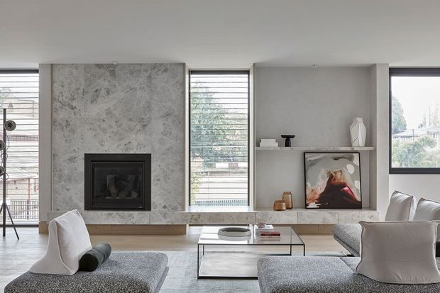 Contemporary living room with a large grey stone fireplace surround and integrated hearth, wide horizontal windows with blinds, built-in open shelving, and low neutral-toned seating arranged around a glass coffee table. The room has a restrained, refined palette of grey, white, and natural stone with abundant daylight.
