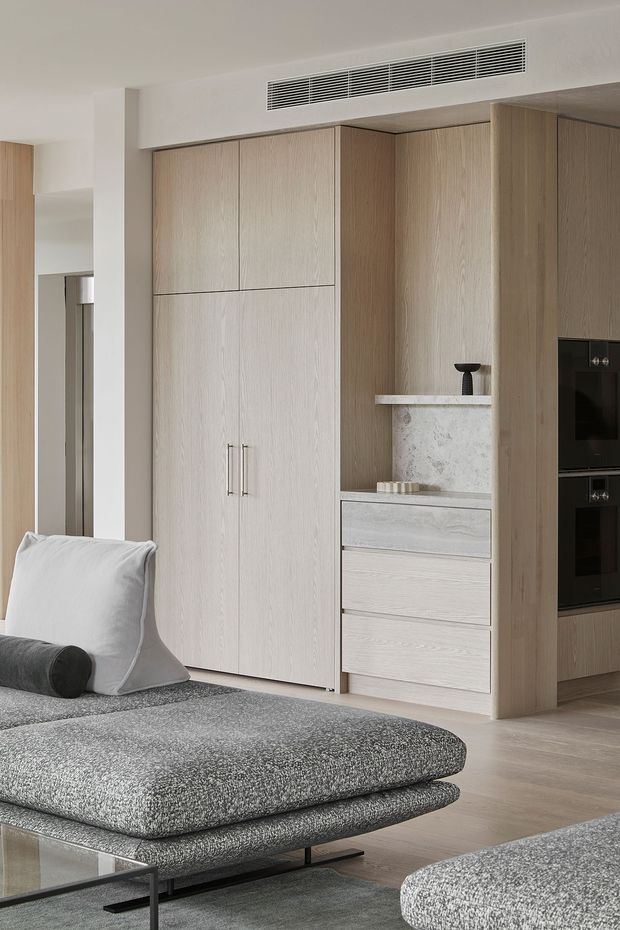 A refined open-plan living area featuring extensive light timber built-in cabinetry, integrated black appliances, a pale stone display niche, and a large textured grey sectional sofa with ottoman seating. The neutral palette and concealed storage create a calm, contemporary interior.