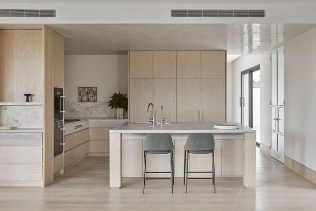 A bright, refined contemporary kitchen viewed from the open living area. The room features a large pale stone island with integrated sink and twin stools, light timber cabinetry extending to the ceiling, pale timber flooring, white walls and ceiling, integrated appliances, and full-height glazing or glazed doors along the right side. The palette is light and neutral with subtle grey-green upholstery and minimal decor.