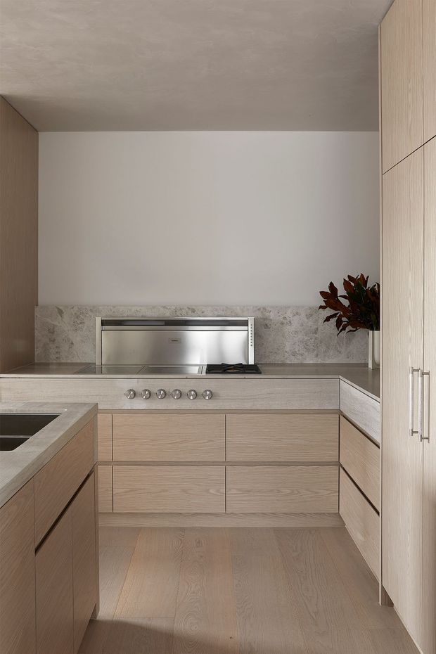 A refined contemporary kitchen with pale timber cabinetry, a matching timber floor, light stone benchtops and a softly patterned stone splashback. The compact U-shaped layout includes an integrated cooktop and oven, island workspace, and tall pantry cabinetry. Neutral tones, concealed storage, and minimal detailing create a calm, understated interior.
