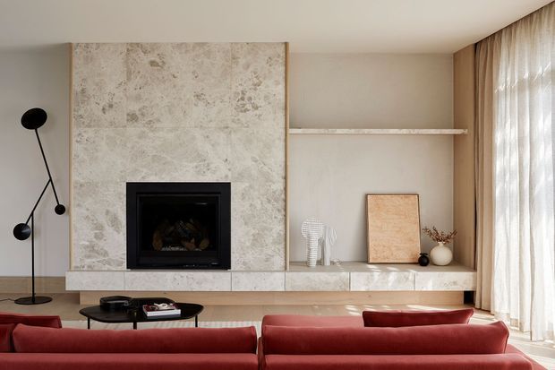 Warm, refined contemporary living room with a large pale stone-clad fireplace wall, integrated open shelving, low stone hearth and built-in cabinetry. Two deep red sofas face a dark oval coffee table, while full-height sheer curtains line the glazed wall. Minimal sculptural decor and a slim black floor lamp complete the calm, curated interior.
