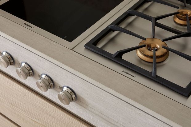 Close-up product-style view of a built-in cooking hob combining a black glass induction zone with a stainless-steel gas cooktop. The gas burners have brass-coloured caps and black cast-iron pan supports, while four metallic control knobs sit along the front on a light wood-grain finished surface.