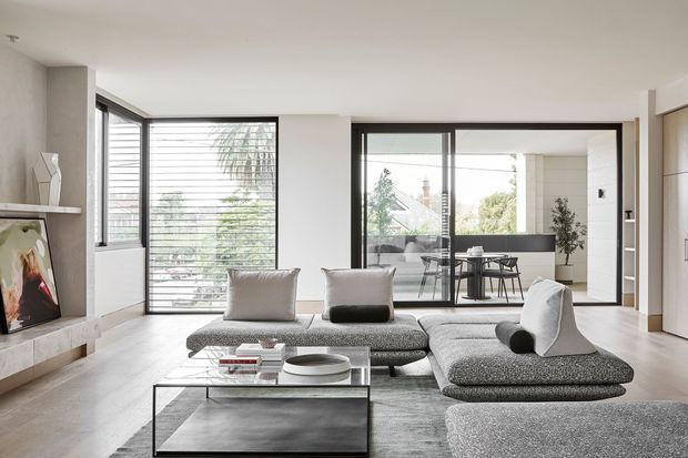 Bright contemporary living room with large floor-to-ceiling windows and black-framed glazing opening toward an outdoor dining area. The room features a low modular grey sectional sofa, glass coffee table, pale timber flooring, a minimalist fireplace wall with artwork, and neutral built-in cabinetry. The interior has a clean, refined, open-plan feel with strong indoor-outdoor connection.