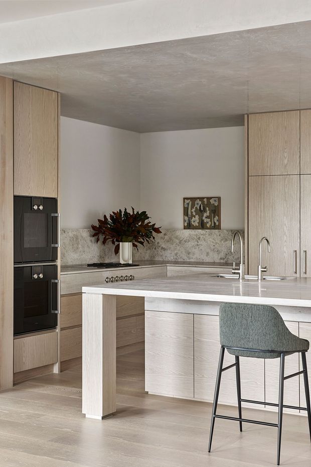 A refined contemporary kitchen with light timber cabinetry, integrated black appliances, a large waterfall-edge island, pale stone benchtops, and a textured stone backsplash. Two curved chrome taps and a single upholstered bar stool sit at the island. The room has a calm, neutral palette with subtle decorative artwork and a broad textured ceiling finish.