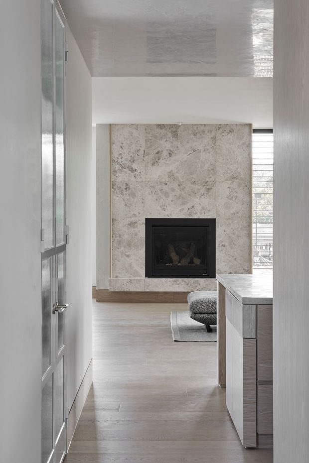 A refined open-plan interior viewed through a glazed door, featuring a large pale natural-stone fireplace surround with a black inset firebox, light timber flooring, white walls and ceiling, and a grey-toned built-in kitchen or cabinetry run in the foreground. A narrow upholstered chair and rug sit near the fireplace, while horizontal window blinds bring in filtered daylight.