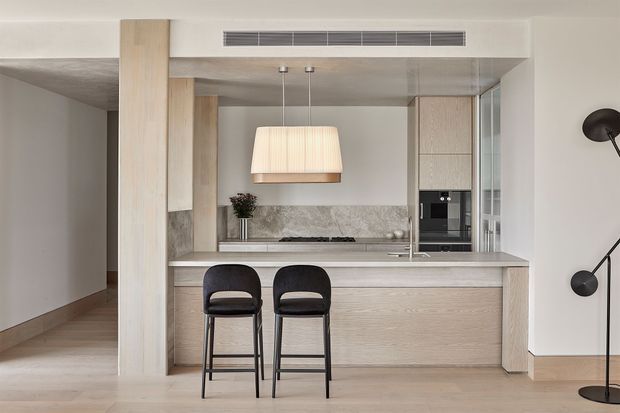 A refined contemporary kitchen viewed from the adjoining living area, featuring a large light timber island with two black bar stools, integrated pale cabinetry, a grey stone splashback and benchtop, built-in appliances, pendant lighting, and a black floor lamp. Pale timber flooring, warm neutral finishes, and clean-lined detailing create a calm, understated interior.