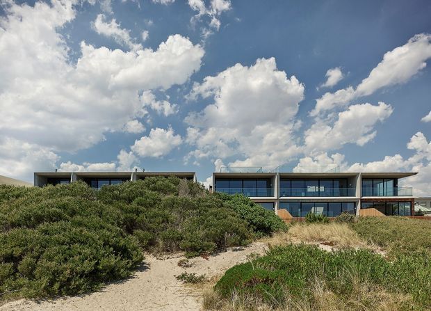 Wide exterior view of a contemporary, low-profile multi-level building set behind coastal sand dunes and native scrub. The facade features extensive dark-framed glazing, horizontal roof planes, balconies or terraces, and sections of warm timber cladding beneath a bright cloud-filled sky.