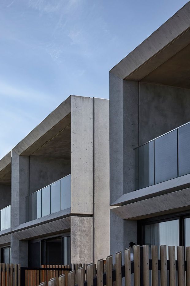 Close-up exterior view of a contemporary multi-level building with strong geometric concrete framing, recessed balconies, clear glass balustrades, dark-framed glazing, and vertical timber privacy fencing. The facade has a raw, minimalist material palette of grey concrete, glass, and timber under a clear sky.