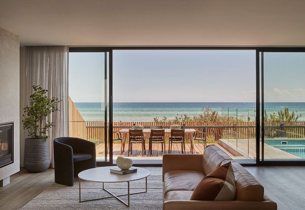 Contemporary open-plan living room facing a beachfront terrace and swimming pool through expansive floor-to-ceiling glazing. The room features a large sectional sofa, sculptural armchair, round coffee table, fireplace, indoor planter, neutral finishes, and a timber outdoor dining deck beyond.