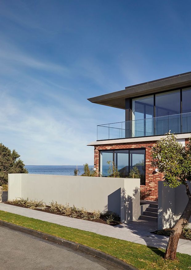 Contemporary multi-storey coastal residence viewed from the street, with a red brick lower facade, extensive dark-framed glazing, a projecting flat roof, and a glass-railed upper balcony. A rendered boundary wall, landscaped front garden, steps, and sidewalk frame the home, with the ocean visible beyond.