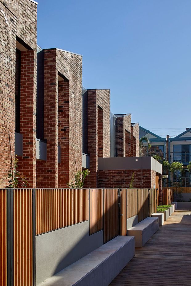 Exterior architectural view of a contemporary multi-level brick development, featuring repeated tall brick volumes, deep recessed openings, timber slatted fencing, concrete seating walls, and a timber boardwalk-style walkway. Clear blue sky and neighbouring pitched-roof homes are visible in the background.