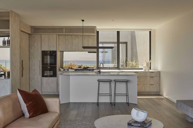 Contemporary open-plan kitchen viewed from the living area, with light timber cabinetry, a large pale island with two black stools, integrated appliances, stone-look flooring, and expansive glazing overlooking a coastal landscape. A linear pendant light and neutral furnishings create a refined, warm interior.