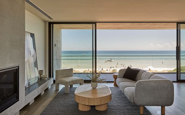 A refined contemporary living room with a broad, floor-to-ceiling glazed opening overlooking a sandy beach and ocean. The room features a minimalist fireplace wall, low built-in media cabinetry, a large rounded upholstered sofa, sculptural lounge chair, round timber coffee table, neutral furnishings, and a textured grey rug.