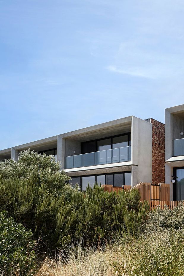Exterior view of a contemporary multi-storey residential building with exposed concrete slab edges, dark-framed glazing, balconies with clear glass balustrades, and sections of red brick wall. Native coastal-style landscaping and grasses occupy the foreground.