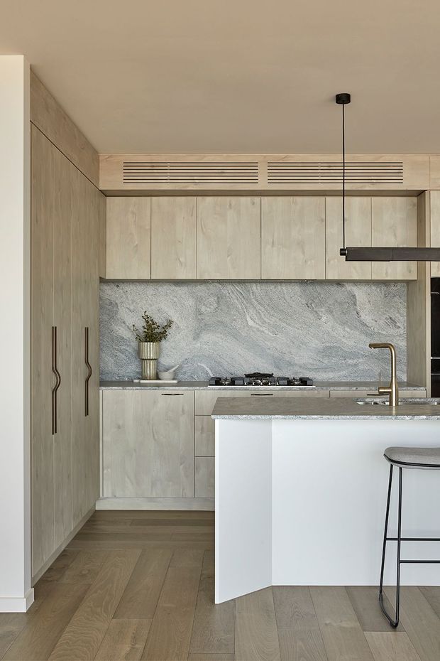 A refined contemporary kitchen with full-height pale timber cabinetry, integrated ventilation grilles, a large pale stone splashback and benchtops, and a white waterfall-edge island. Warm timber flooring, a slim black pendant light, and a brass-toned tap create a restrained, neutral palette.