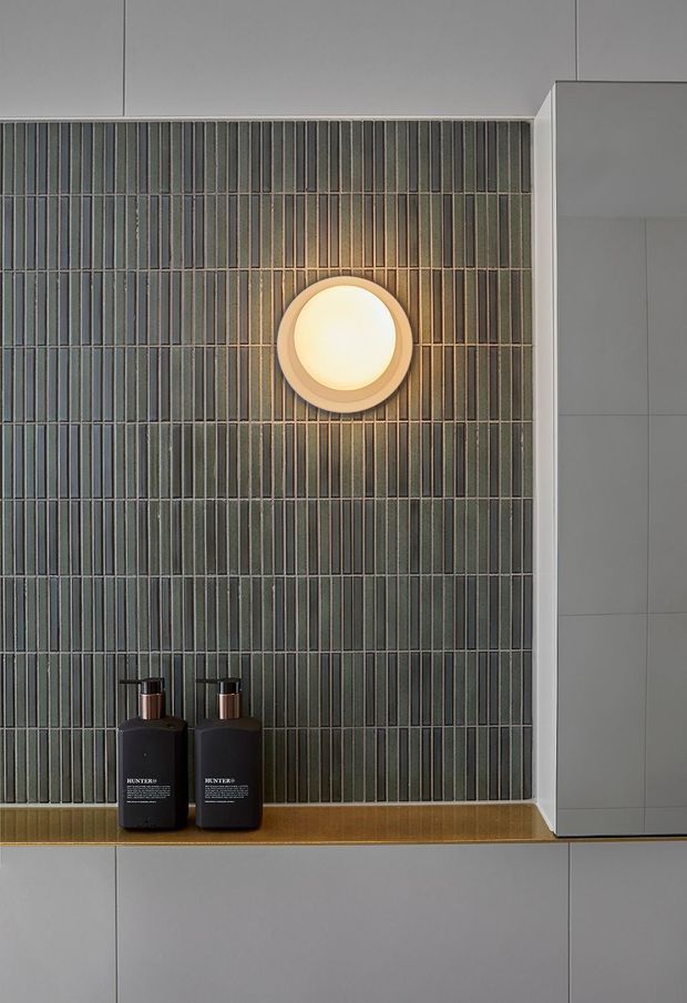 Close-up of a refined bathroom vanity wall featuring dark green-grey vertically ribbed ceramic tile, pale grey built-in cabinetry, a warm round wall light, and a narrow timber-toned shelf with two black pump bottles. The composition is minimalist and carefully detailed, with warm light contrasting against cool tiled and cabinetry surfaces.