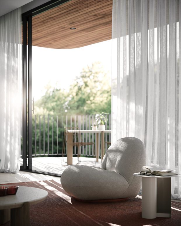Bright contemporary sitting area opening onto a balcony through full-height black-framed glazing. Sheer white curtains soften the sunlight, while a sculptural cream lounge chair, low round coffee table, warm timber ceiling lining, and deep reddish-brown rug create a refined, inviting interior.