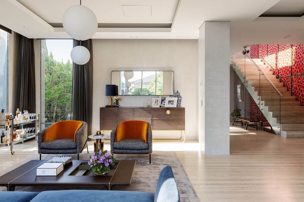 A refined contemporary living room with floor-to-ceiling glazing, dark curtains, two patterned lounge chairs, a low dark coffee table, and a blue sofa in the foreground. A timber sideboard and mirror sit against the rear wall, while an open stairway with a striking red patterned feature wall is visible to the right. Large spherical pendant lights and a recessed ceiling add architectural detail.
