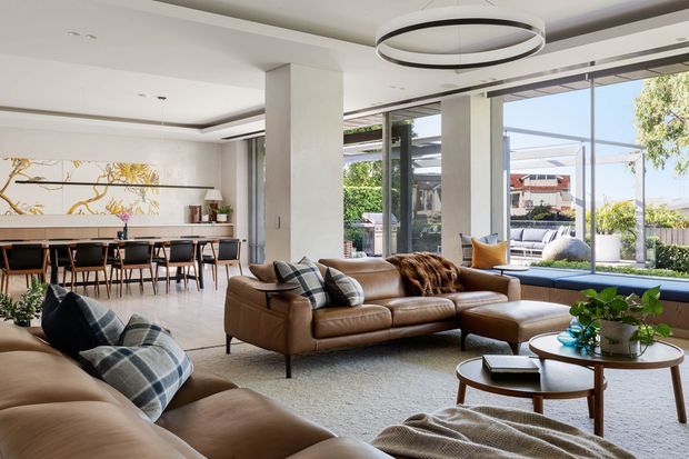 Bright, spacious open-plan living and dining area with large floor-to-ceiling glazing opening to a landscaped outdoor terrace. The room features tan leather sofas, a low timber coffee table, a long dining table with upholstered chairs, a neutral rug, layered cushions, contemporary circular pendant lighting, and built-in window seating. Clean-lined architecture and abundant natural light create a refined, inviting atmosphere.