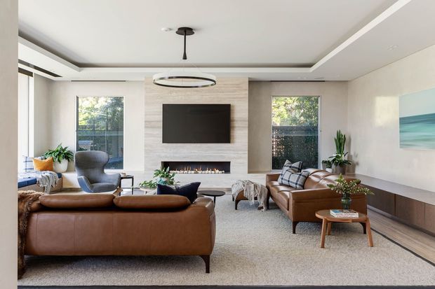 A refined contemporary living room with a large brown leather sectional sofa, matching leather seating, a central wall-mounted television, and a linear fireplace set into a light stone feature wall. The room has a layered tray ceiling, circular pendant light, neutral carpet, timber-toned built-in cabinetry, indoor plants, and large windows overlooking greenery.