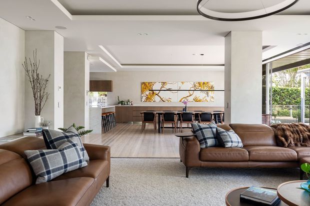 Bright, open-plan contemporary living and dining area with brown leather sofas, plaid cushions, a large textured rug, and a sculptural circular pendant light. The space opens into a dining area and kitchen with timber cabinetry, a long island or dining counter, light timber flooring, white walls, and expansive glazing overlooking greenery.