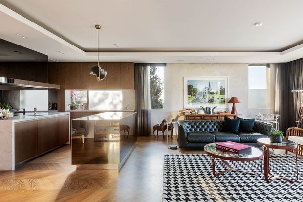 A sophisticated open-plan living, dining and kitchen interior with dark timber cabinetry, a central stone island, parquet timber flooring, a black leather sofa, patterned area rug and mid-century-inspired furniture. Large windows provide abundant natural light, while a recessed ceiling detail and pendant light add architectural interest.