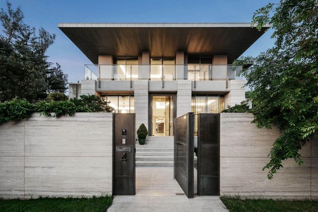 Front exterior view of a refined contemporary multi-level residence, featuring a broad flat projecting roof, exposed concrete walls and columns, extensive upper-level glazing with clear balustrades, and a formal entry approached by broad concrete steps. Dark metal pedestrian gates and dense landscaping frame the facade.