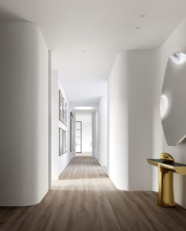 A bright, minimalist residential hallway with sculptural curved white plaster walls, pale timber flooring, recessed ceiling lights, framed artwork, and a small gold console table with decorative objects. The corridor leads toward a glazed door and is defined by soft organic forms and abundant natural light.