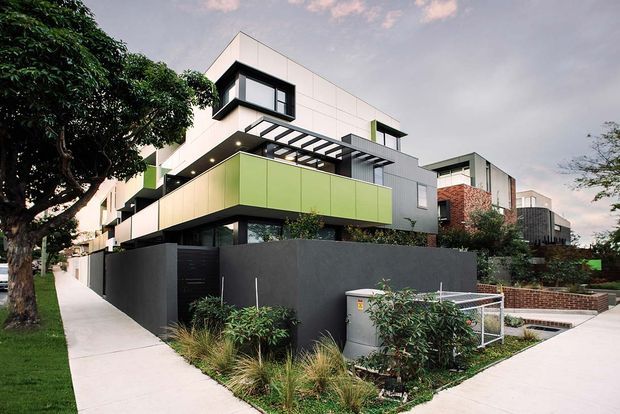 Street-level view of a contemporary multi-storey residential building with crisp white and grey rendered façades, projecting box forms, lime-green cladding panels, dark window frames, and a pergola-like balcony structure. The property is enclosed by a dark masonry boundary wall and landscaped front garden, with a pedestrian footpath and driveway visible in the foreground.