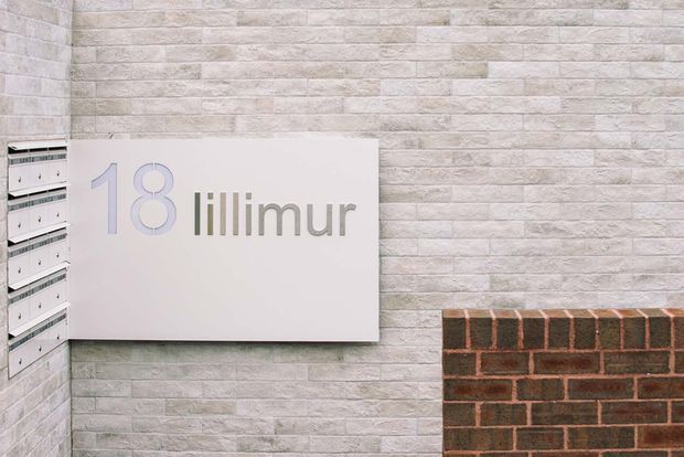 Close-up of a contemporary building entrance or facade featuring a pale textured masonry wall, a large white address and building-name sign reading “18 lillimur,” recessed mailboxes at the left, and a contrasting dark brick base at the lower right.