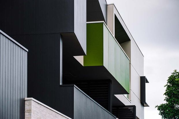 A close-up exterior view of a contemporary multi-level building with bold geometric forms, dark vertical cladding, pale panels, a vivid green recessed facade section, projecting balconies or overhangs, and layered privacy screens. A rendered masonry wall and partial landscaping are visible at the lower edge.