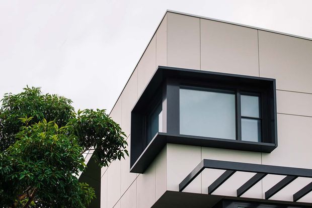 Close-up exterior view of a contemporary multi-level building with pale large-format facade panels, bold dark window surrounds, projecting box window, and a dark-framed canopy. A leafy tree partially screens the lower left side.