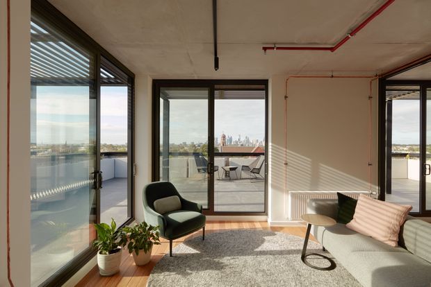 Bright contemporary living room with floor-to-ceiling glazed doors opening onto a terrace or balcony. The room features exposed concrete ceilings, visible red service piping, timber flooring, a large textured rug, a pale sofa, sculptural armchair, round side table, indoor plants, and expansive urban views.