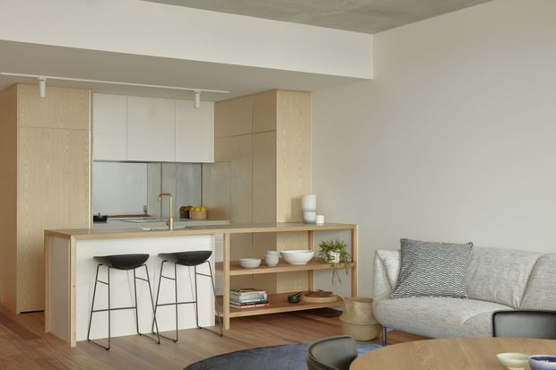 A bright open-plan contemporary living space combining a compact kitchen, island breakfast bar, and living area. Pale timber cabinetry and open shelving frame the kitchen, with a stainless-steel or reflective splashback, white cabinetry, and brass-toned tapware. Two black stools sit at the island, while the adjacent lounge includes a light grey sofa, patterned cushion, round coffee table, and neutral decor. The room has a clean, restrained palette and a low concrete ceiling beam.
