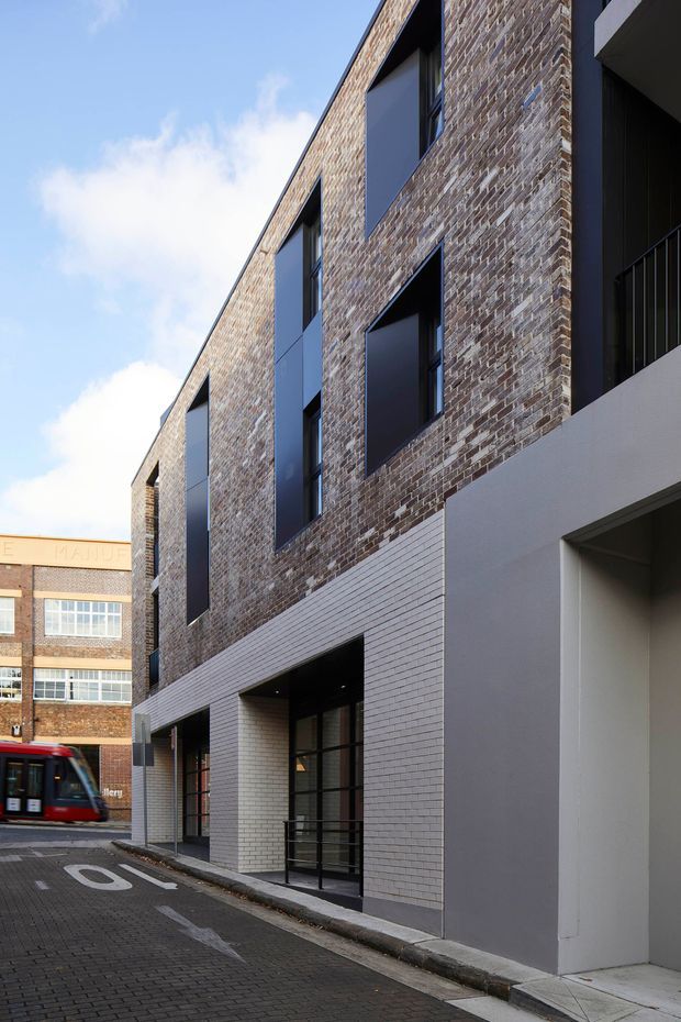 Street-level view of a contemporary multi-storey urban building with a predominantly exposed brown brick facade, dark recessed window openings and frames, light grey brick at ground level, and smooth grey rendered sections. The building fronts a narrow paved lane with an adjacent older brick commercial building and a partially visible tram in the background.