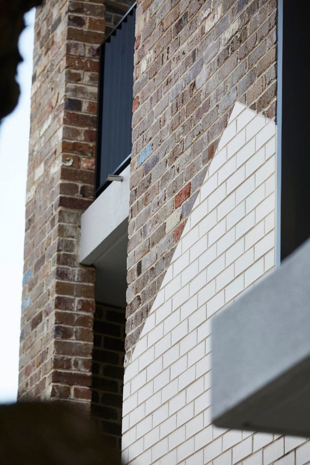 Close-up architectural exterior detail showing a contemporary facade composed of weathered red-brown brickwork and a contrasting field of small white rectangular tiles. Recessed openings, dark metal screening, and projecting grey concrete elements create a layered, tactile composition.