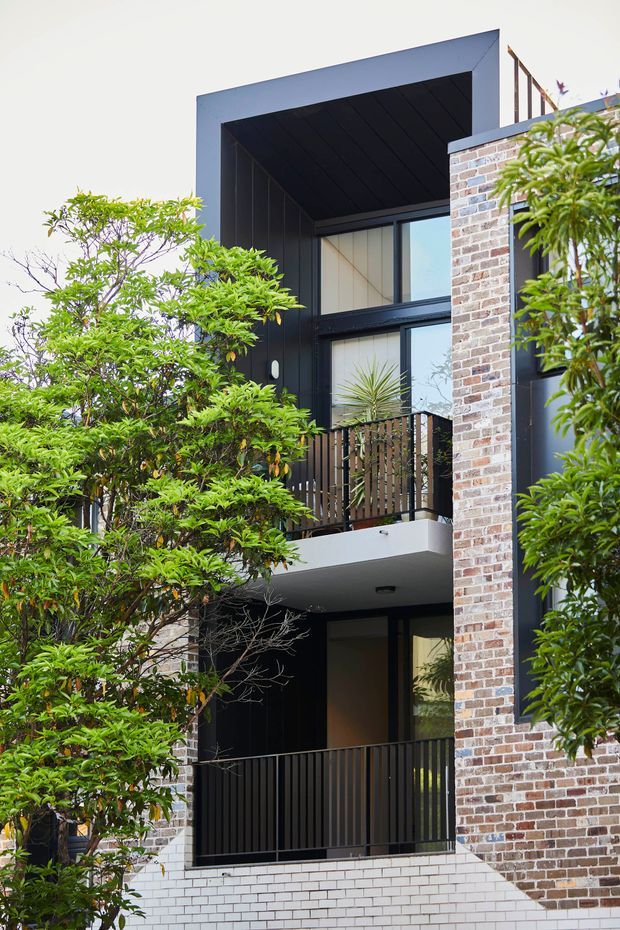 Vertical exterior view of a contemporary multi-storey building partially screened by leafy landscaping. The facade combines dark cladding and deep recessed balconies with black metal balustrades, large glazing, and contrasting exposed brickwork in varied pale and reddish tones. A projecting upper-level roof or frame creates a strong geometric portal around the windows and balcony.
