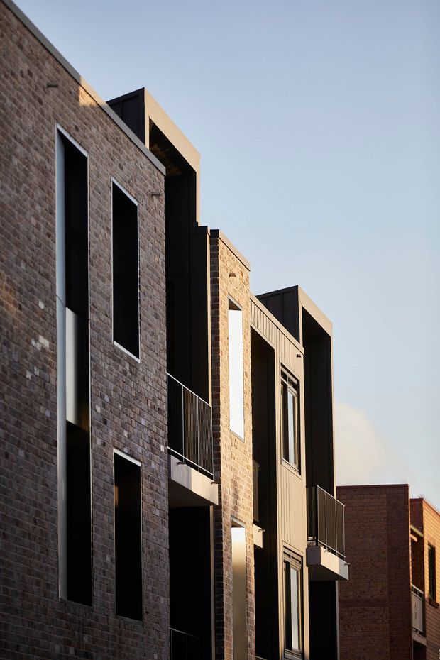 A close-up exterior view of a contemporary multi-storey residential building. The facade combines warm brown brickwork with dark metal-clad vertical volumes, deep-set windows, projecting balconies, and slim black balustrades. Strong late-afternoon sunlight highlights the textured masonry and geometric massing against a clear sky.