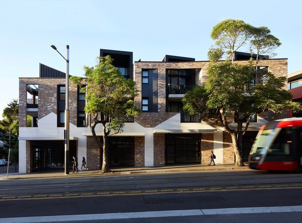Street-facing view of a contemporary multi-storey urban residential or mixed-use building with a warm brick facade, dark-framed windows, projecting balconies, recessed entrances, and white textured geometric facade elements. Mature street trees, tram tracks, and a passing red tram establish a dense public transport-oriented city setting.