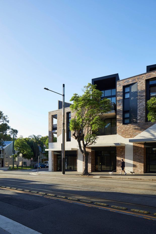 Street-level exterior photograph of a contemporary multi-storey urban residential or mixed-use building. The facade combines warm brown brickwork with pale masonry, dark window frames, recessed balconies, projecting box forms, and ground-floor glazing. A tree-lined sidewalk, street, tram or traffic-calming infrastructure, and neighbouring buildings are visible under a clear blue sky.