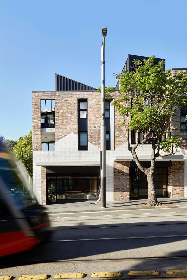 Street-facing multi-storey contemporary brick building with a mixed brick and white-painted masonry facade, tall narrow windows, recessed balconies, dark metal roof forms, and ground-level glazed commercial or entry spaces. A mature street tree, footpath, road, tram or traffic infrastructure, and a blurred passing vehicle are visible in the foreground under clear blue sky.