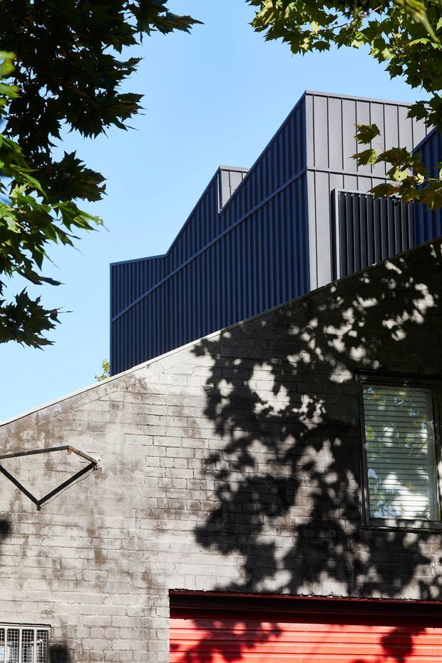 Exterior architectural photograph of a compact urban building viewed from below, framed by leafy tree branches against a clear blue sky. The lower structure has a weathered grey concrete-block wall with a small louvered window and a bright red roller garage door, while the upper level features strongly ribbed dark blue and light grey metal cladding with stepped, angular roof forms.