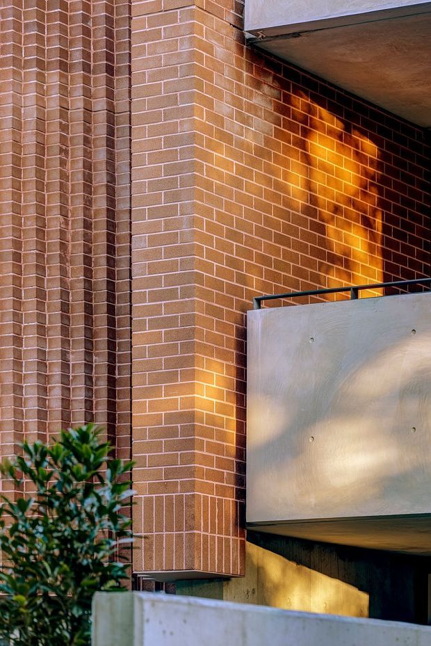 Close-up exterior architectural view of a multi-level brick building, featuring textured projecting brick piers, warm accent lighting washing across the brickwork, a smooth metal-clad projecting balcony or facade element, dark metal railing, and foreground landscaping. The composition emphasises material texture, structural depth, and warm evening illumination.