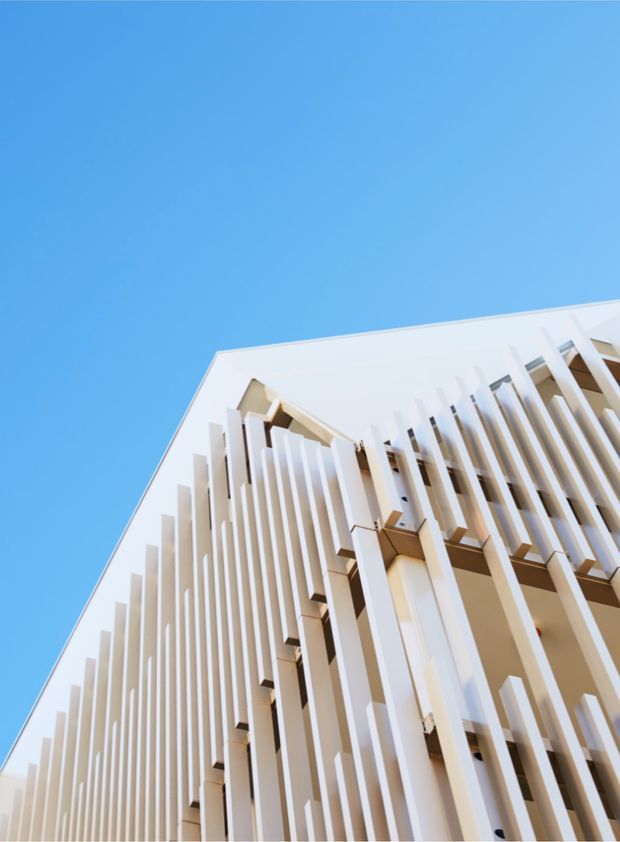 Upward-looking architectural photograph of a contemporary multi-storey building facade under a clear blue sky. The bright white facade is defined by dense vertical fins or sun-shading screens, with recessed balconies or glazing visible behind them. The perspective emphasizes the building's height and geometric repetition.