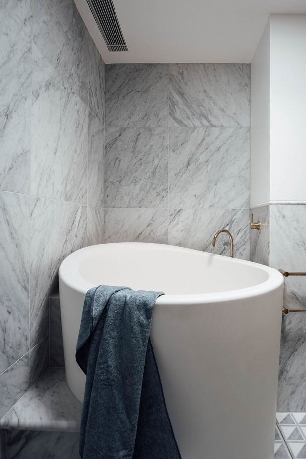 A compact, refined bathroom featuring a freestanding white bathtub surrounded by large grey-white veined marble tiles. Brushed brass plumbing fixtures and a dark blue towel provide understated contrast, while a linear ceiling air vent and patterned floor tile are also visible.