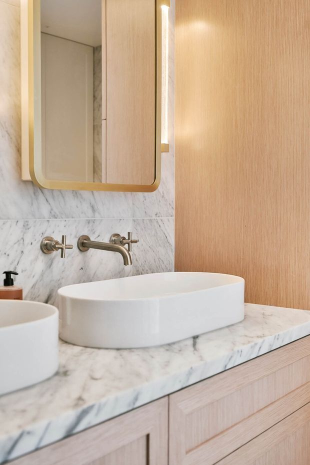 Close-up of a refined bathroom vanity featuring a white vessel basin, a marble countertop and wall finish, brushed metal wall-mounted tapware, light timber-grain cabinetry, and a rounded rectangular mirror with a warm metallic frame. Soft integrated lighting and warm timber tones create a calm, luxurious feel.