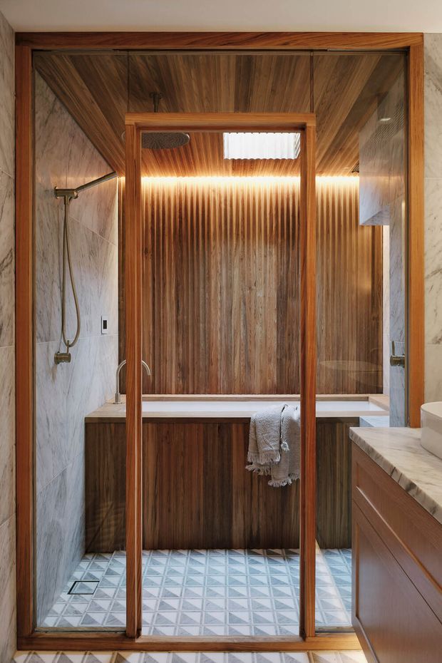 A refined bathroom featuring a walk-in shower enclosed by a timber-framed glass screen, a built-in bathtub with vertically grained dark timber wall panelling, and a timber vanity with a light marble benchtop. Pale marble-look wall tiles and patterned geometric floor tiles add contrast, while integrated linear lighting creates a warm glow.