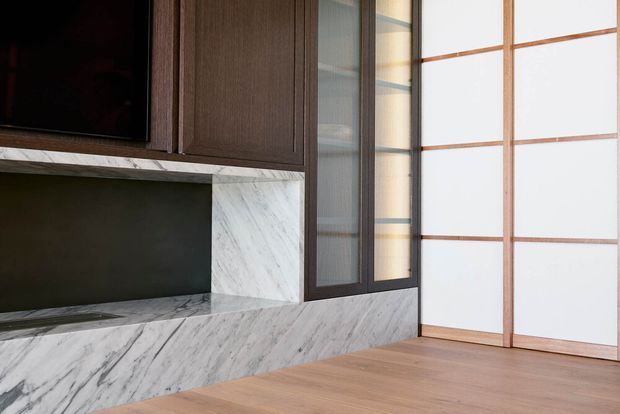 A refined contemporary living room media wall featuring dark timber cabinetry, a recessed dark display niche, and a substantial veined marble surround and hearth. A large black television is partially visible above, while timber flooring and a translucent timber-framed sliding screen or partition occupy the right side of the image.