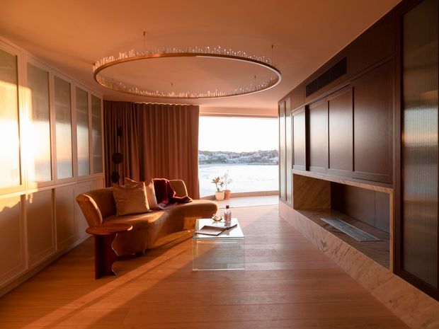 Warm, refined living room overlooking a body of water through large glazing. The room features rich timber flooring, dark wood wall panelling and built-in cabinetry, a sculptural curved sofa, a glass coffee table, heavy brown curtains, and a large circular pendant light with crystal-like detailing. The palette is dominated by warm wood, brown, and amber tones, with strong evening or sunset light.